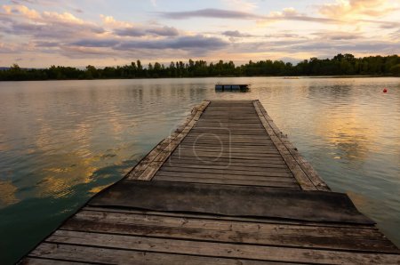 Foto de Vista pacífica del atardecer en un lago del centro de ocio de Les Etangs, en Saix, al sur de Francia, con un embarcadero de madera flotando en el agua y los bosques de la reserva natural en el fondo - Imagen libre de derechos
