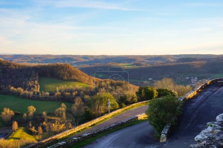 Téléchargez les photos : Superbe panorama sur un plateau verdoyant vu depuis les remparts médiévaux de Puycelsi, une ville fortifiée du sud de la France, au-dessus d'une route serpentine de campagne qui serpente autour de la montagne - en image libre de droit