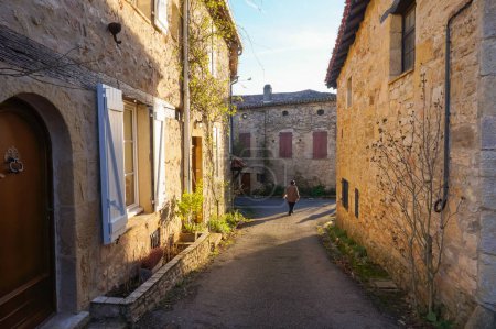 Téléchargez les photos : Puycelsi, France - Décembre 2018 - Une étroite rue médiévale bordée de vieilles maisons typiques avec des murs rocheux avec du lierre grimpant sur la pierre ; à l'arrière, un homme marche seul dans l'allée - en image libre de droit
