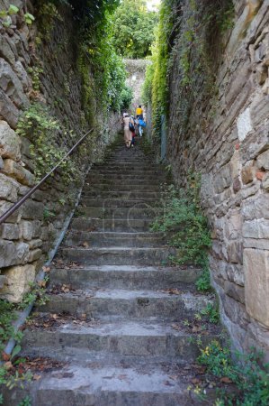 Foto de Cordes-sur-Ciel, Francia - Agosto de 2021 - Una impresionante escalera al aire libre en el pueblo medieval, flanqueada por dos enormes paredes con hiedra pegada a las piedras, y turistas subiendo a la cima - Imagen libre de derechos