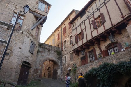 Photo for Cordes-sur-Ciel, France - Aug. 2021 - An old, typical street in the medieval village, with an arched gate, traditional town buildings and a half-timbered, corbelled house with cantilevered parts - Royalty Free Image