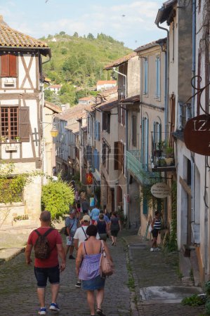 Foto de Cordes-sur-Ciel, Francia - Agosto de 2021 - Los turistas que visitan el pueblo medieval en "Grand-Rue de l 'Horloge", una calle inclinada bordeada de negocios tradicionales locales, y dominada por las colinas de Sgala - Imagen libre de derechos