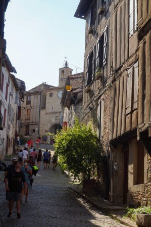 Foto de Cordes-sur-Ciel, Francia - Agosto de 2021 - Turistas recorriendo una calle empedrada inclinada bordeada de casas antiguas típicas, en el pintoresco pueblo medieval; hay una puerta fortificada en la subida - Imagen libre de derechos
