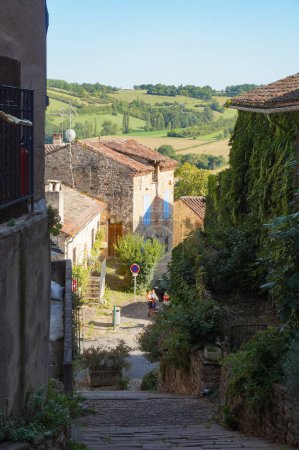 Photo for Cordes-sur-Ciel, France - Aug. 2021 - Verdant, hilly landscape seen from a sloping, cobbled street lined with old, traditional houses, in Cordes, a picturesque medieval village in Midi-Pyrenes - Royalty Free Image