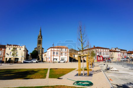 Photo for Villefranche d'Albigeois, France - May 2020 - Refurbished main square of the village, planted with young trees, and old traditional brick houses dominated by the bell tower of the neo-Gothic church - Royalty Free Image