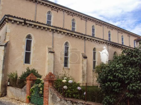 Foto de Fachada lateral de la iglesia neogótica de Villefranche d 'Albigeois, un pueblo del sur de Francia; las paredes están atravesadas por vidrieras y dominan un jardín con una estatua de la Virgen - Imagen libre de derechos