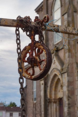 Photo for Ancient rusty pulley and chain of an old well, in the middle of the square that is in front of the neogothic church of Villefranche d'Albigeois, a countryside village in the South of France - Royalty Free Image