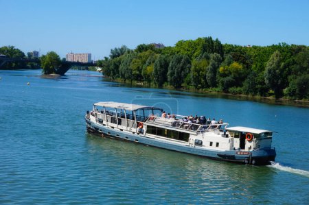 Foto de Toulouse, Francia - Julio 2020 - Una barcaza de cruceros embarcando a los turistas en la cubierta para un recorrido turístico por el río Garona, navega frente al frondoso parque de la Prairie des Filtres, en la orilla - Imagen libre de derechos