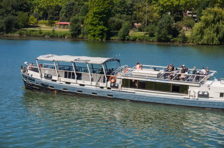Foto de Toulouse, Francia - Julio 2020 - Una barcaza de cruceros embarcando a los turistas en la cubierta para un recorrido turístico por el río Garona, navega frente al frondoso parque de la Prairie des Filtres, en la orilla - Imagen libre de derechos