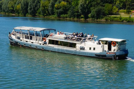 Photo for Toulouse, France - July 2020 - A cruise barge embarking tourists on the deck for a sighseeing tour on the River Garonne, navigates in front of the leafy park of the Prairie des Filtres, on the bank - Royalty Free Image