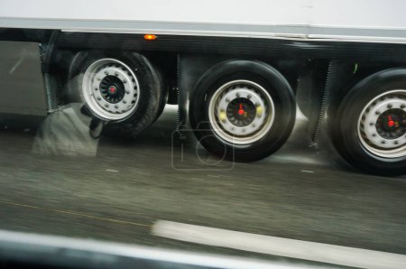 Photo for Motion-blurred photo of the wheels in rapid rotation of a trailer truck driving at high speed by rainy weather on a wet road and projecting splash and drops of water on the asphalt of the motorway - Royalty Free Image