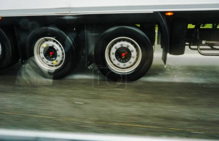 Téléchargez les photos : Photo floue des roues en rotation rapide d'une semi-remorque conduisant à grande vitesse par temps pluvieux sur une route mouillée et projetant éclaboussures et gouttes d'eau sur l'asphalte de l'autoroute - en image libre de droit