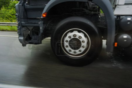 Foto de Foto borrosa por movimiento de las ruedas en rotación rápida de un remolque tractor conduciendo a alta velocidad por el clima lluvioso en una carretera mojada y proyectando salpicaduras y gotas de agua en el asfalto de la autopista - Imagen libre de derechos