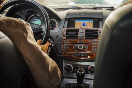 Téléchargez les photos : Un automobiliste conduisant sa voiture et tenant le volant, vu de derrière, avec vue sur le tableau de bord et la console centrale avec l'écran GPS ; être derrière le volant pour un road trip - en image libre de droit
