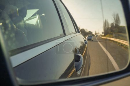 Photo for Looking behind in the rearview mirror of the car, reflecting the handle of the door, the chrome bar and bodyshell of the car, and the motorway with a car, road marking and guardrail in the blur - Royalty Free Image