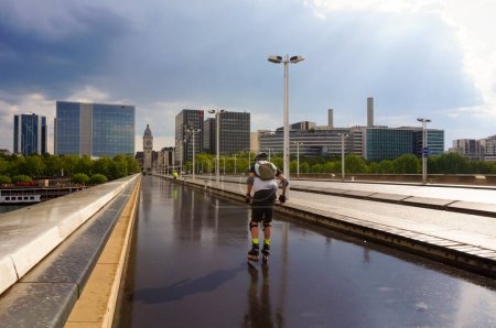 Téléchargez les photos : Paris, France - Mai 2021 - Un homme roule sur l'asphalte mouillé, sur le trottoir du pont Charles de Gaulle, qui traverse la Seine ; de loin, la tour de l'horloge de la gare de Lyon - en image libre de droit