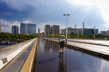 Foto de París, Francia - Mayo 2021 - Un hombre patina sobre asfalto mojado, en la acera del puente Charles de Gaulle, que cruza el río Sena; a lo lejos, la torre del reloj de la estación de tren Gare de Lyon - Imagen libre de derechos