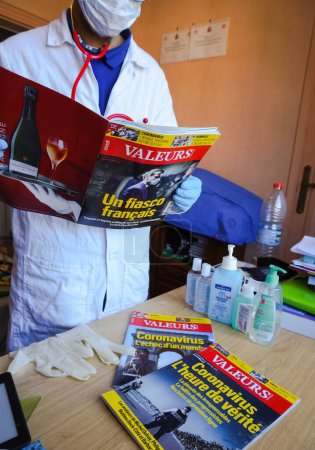 Photo for Albi, France -March 2020- Hydroalcoholic gel, gloves and issues of the French newspaper Valeurs Actuelles about the coronavirus read by a physician wearing a white blouse and a mask (staged photo) - Royalty Free Image