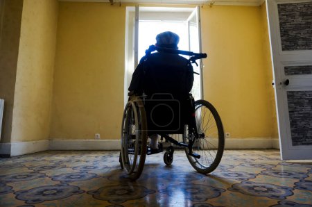 Téléchargez les photos : Tarn, France - Mai 2020 - Un vieil homme handicapé avec un béret assis sur un fauteuil roulant, regardant par une fenêtre ouverte ouvrant sur le jardin ensoleillé, seul dans une vaste pièce vide dans une ancienne maison de campagne - en image libre de droit