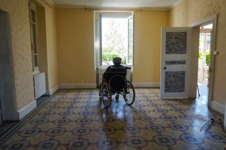 Foto de Tarn, Francia - Mayo 2020 - Un anciano discapacitado con una boina se sienta en una silla de ruedas, mirando a través de una ventana abierta que da al soleado jardín, solo en una vasta habitación vacía en una antigua casa de campo - Imagen libre de derechos