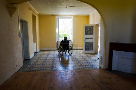 Téléchargez les photos : Albi, France - Mai 2020 - Un vieil homme handicapé avec un béret assis sur un fauteuil roulant, regardant par une fenêtre ouverte ouvrant sur le jardin ensoleillé, seul dans une vaste pièce vide dans une ancienne maison de campagne - en image libre de droit