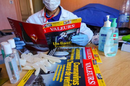 Photo for Albi, France -April 2020- Hydroalcoholic gel, gloves and issues of the French newspaper Valeurs Actuelles about the coronavirus read by a physician wearing a white blouse and a mask (staged photo) - Royalty Free Image