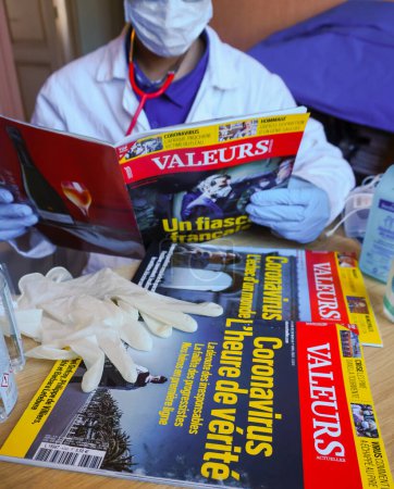 Téléchargez les photos : Albi, France - Avril 2020- Gel hydroalcoolique, gants et numéros du journal français Valeurs Actuelles sur le coronavirus lus par un médecin portant une blouse blanche et un masque (photo mise en scène) - en image libre de droit