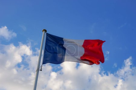 The three-colored, blue, white and red national flag of France, flown on top of a white metal pole, and undulating in the wind, with a beautiful blue sky in the background and some white clouds