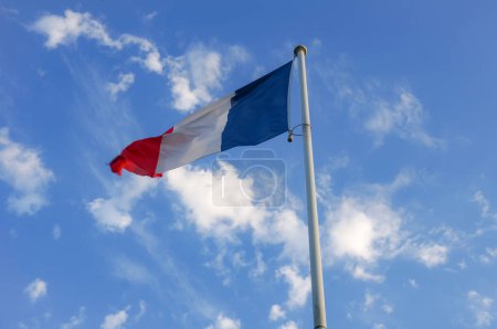 Téléchargez les photos : Le drapeau national trois couleurs, bleu, blanc et rouge de la France, flotté au sommet d'un poteau métallique blanc, et ondulant dans le vent, avec un beau ciel bleu en arrière-plan et quelques nuages blancs - en image libre de droit