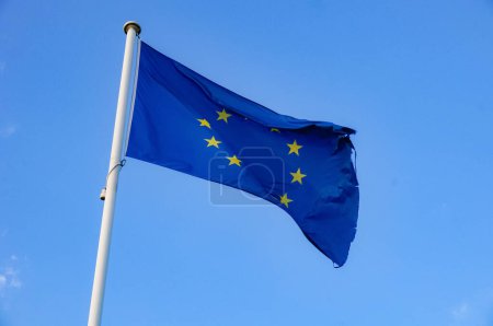 Téléchargez les photos : Drapeau endommagé et légèrement déchiré de l'Union européenne, avec les douze étoiles sur fond bleu, agitant dans les airs, dans un ciel bleu clair à Sax, Sud de la France, au sommet d'un mât métallique blanc - en image libre de droit