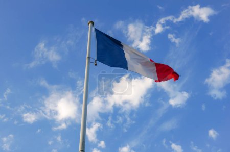 Téléchargez les photos : Le drapeau national trois couleurs, bleu, blanc et rouge de la France, flotté au sommet d'un poteau métallique blanc, et ondulant dans le vent, avec un beau ciel bleu en arrière-plan et quelques nuages blancs - en image libre de droit