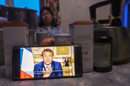 Téléchargez les photos : Albi, France - 13 avril 2020 - Une Française écoute avec anxiété l'allocution d'Emmanuel Macron, diffusée en direct sur un téléphone par la télévision publique, à propos du confinement dû au coronavirus, lors d'un dîner - en image libre de droit
