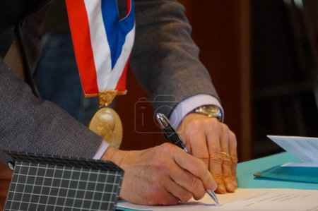 Téléchargez les photos : Cimier, France - Novembre 2021 - Le maire Herve Mariton, portant une médaille officielle dorée avec une ceinture bleue, blanche et rouge, et tenant un stylo, signe des documents officiels à la mairie lors d'une cérémonie de mariage - en image libre de droit