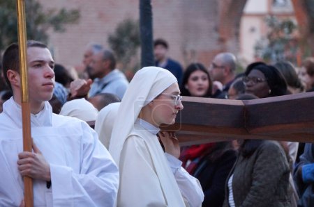 Téléchargez les photos : Toulouse, France - Mars 2023 - Une jeune religieuse catholique porte une croix de bois, tandis qu'un jeune moine porte une bougie, portant tous deux des soutènements blancs, lors d'une procession religieuse pour célébrer l'Annonciation - en image libre de droit