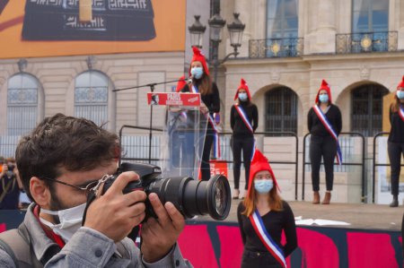 Foto de París, Francia - 10 de octubre de 2020 - Un fotógrafo de prensa en acción en la manifestación de Marchons Enfants contra el proyecto de ley de bioética, mientras una "Marianne" del Manif pour Tous da un discurso sobre el escenario - Imagen libre de derechos