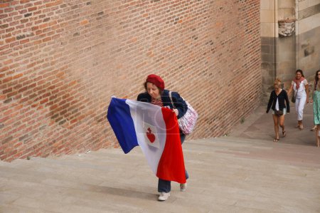Téléchargez les photos : Albi, France - 24 juillet 2021 - Une femme âgée monte les escaliers de la Cathédrale, tenant un drapeau français marqué d'un Sacré-Cœur ("Coeur de Vendee"), en marge d'une manifestation anti-santé - en image libre de droit