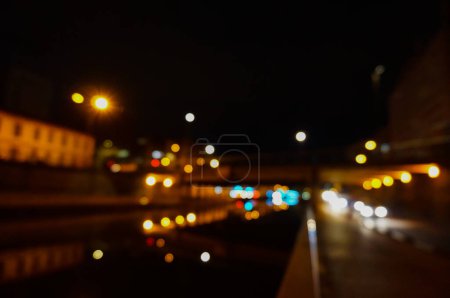 Photo for Blur image of night lights in the street, with reflections from headlights or lampposts in the water of the canal, on the road and under the bridge ; there are yellow, white, blue and red lights - Royalty Free Image