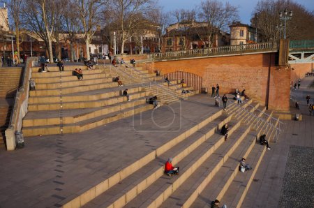 Photo for Toulouse, France - Feb. 2020 - Tourists and locals enjoying the sunset on the stairs of the popular and famous Saint-Pierre Square, on the embankment of the River Garonne, next to the metal bridge - Royalty Free Image