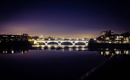 Photo for Night view of lights from the windows of the residential buildings of Bazacle Promenade Dike, in Toulouse, France, and Pont des Catalans Bridge crossing the river Garonne with reflection in the water - Royalty Free Image