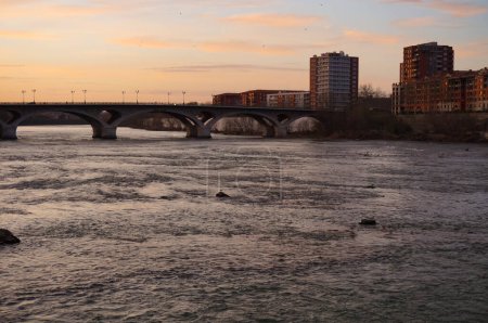 Téléchargez les photos : Coucher de soleil sur la Garonne et silhouette de bâtiments résidentiels au bord de la rivière, dans le quartier du Bazacle Dike à Toulouse, France, avec le pont voûté du Pont des Catalans - en image libre de droit