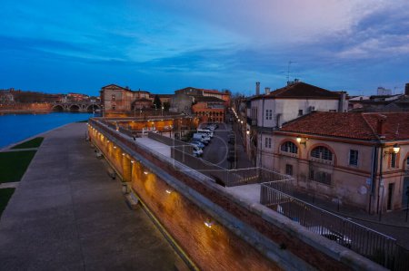 Téléchargez les photos : Vue de nuit de vieilles maisons de ville en briques traditionnelles à Saint-Cyprien, derrière la passerelle construite sur le brise-lames illuminé de l'ancien port fluvial de Viguerie, au bord de la Garonne à Toulouse, France - en image libre de droit