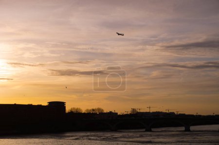 Foto de Vista al atardecer del río Garona que fluye bajo el puente Pont des Catalans en Toulouse, Francia, con siluetas de edificios y grúas, y de un avión despegando sobre la ciudad en un cielo amarillo - Imagen libre de derechos