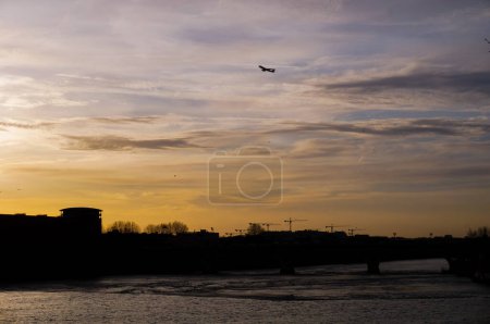 Foto de Vista al atardecer del río Garona que fluye bajo el puente Pont des Catalans en Toulouse, Francia, con siluetas de edificios y grúas, y de un avión despegando sobre la ciudad en un cielo amarillo - Imagen libre de derechos