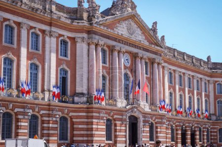 Photo for The Capitole, city hall of Toulouse, France, with its monumental brick classic facade and marble columns brick, richly adorned with French flags plus European and Occitan flags hanged on the balcony - Royalty Free Image