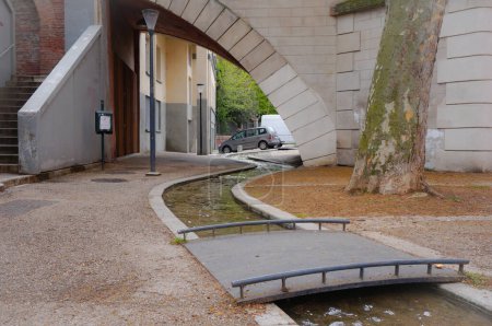Foto de Pequeño y encantador arroyo serpenteante que fluye bajo el arco flanqueado por la escalera del puente Pont de Tounis, atravesado por una pasarela, y el tronco de un platano, en Avenue de la Garonnette, Toulouse, Francia - Imagen libre de derechos