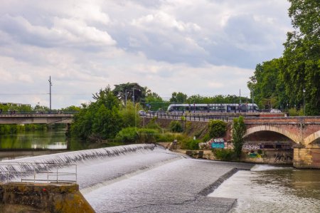 Foto de Toulouse, Suroeste de Francia - Julio 2020 - El río Garona que fluye sobre la presa del barrio de Tounis, con un tren del tranvía que cruza el puente de Saint-Michel en el fondo - Imagen libre de derechos
