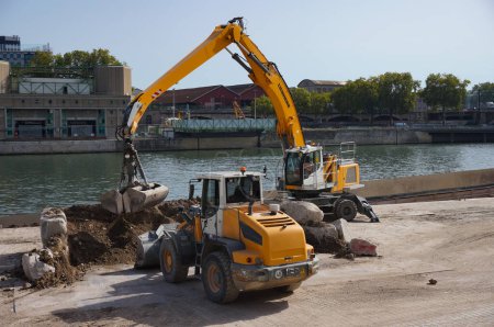 Foto de París, Francia - 6 de septiembre de 2023 - cargador Komatsu amarillo y excavadora Liebherr, con un brazo hidráulico extendido, en catión en un muelle junto al agua en Port de Tolbiac, un puerto industrial junto al río Sena - Imagen libre de derechos