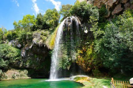 Foto de La conocida cascada, que fluye de un promontorio rocoso a una piscina natural que da reflejos verdes al agua, en Salles-la-Source, un pueblo rural en el departamento de Aveyron en el sur de Francia - Imagen libre de derechos