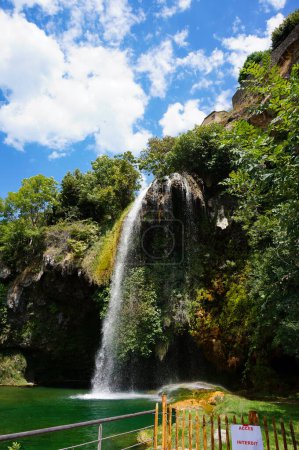 Foto de La conocida cascada, que fluye de un promontorio rocoso a una piscina natural que da reflejos verdes al agua, en Salles-la-Source, un pueblo rural en el departamento de Aveyron en el sur de Francia - Imagen libre de derechos