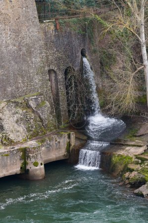Foto de Saint-Jury, Francia - 28 de enero de 2024 - El agua que fluye hacia el río Tarn desde un conducto dispuesto en la pared de una fábrica metalúrgica histórica construida en el terraplén, en Saut du Sabo Cataract - Imagen libre de derechos
